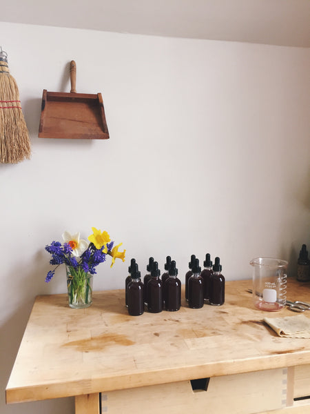 Wooden workspace with fresh flowers, tinctures, straw broom and measuring beaker.